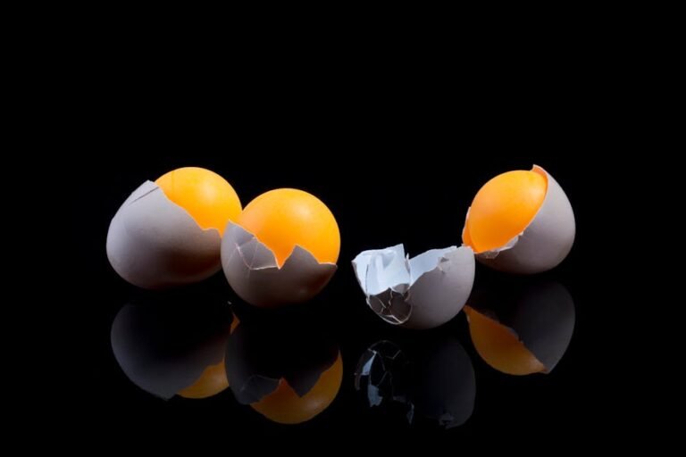 Two cracked eggs with bright yolks on a reflective black surface, showcasing culinary ingredients.