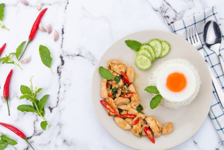 Thai meal featuring basil chicken, chili peppers, rice, and a fried egg on a marble background.
