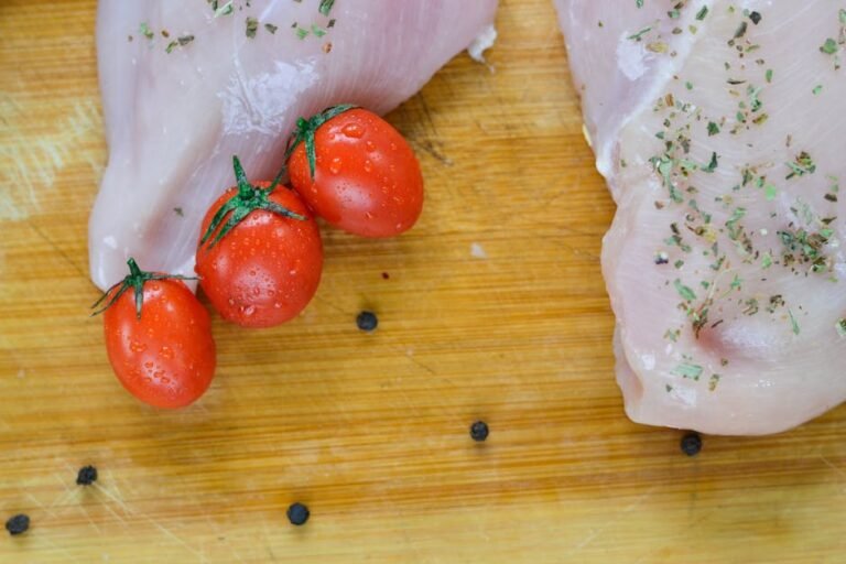 Close-up of raw chicken breast garnished with herbs, paired with fresh cherry tomatoes on a wooden surface.